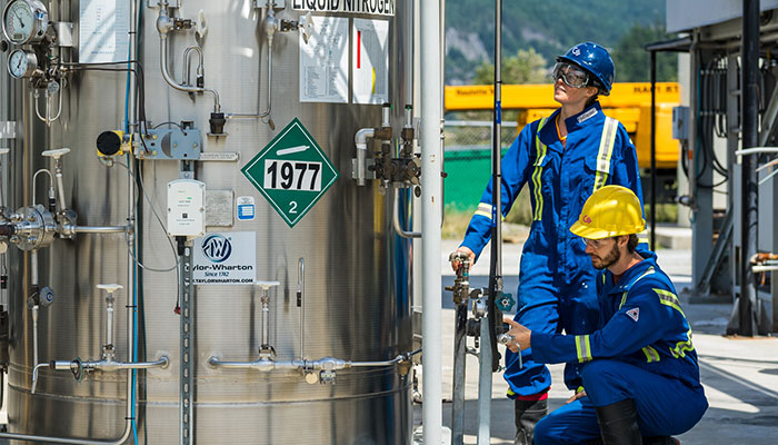 Two Carbon Engineering employees in blue coveralls take measurements outside at a direct air capture facility with mountains in the background and liquid nitrogen in the foreground.