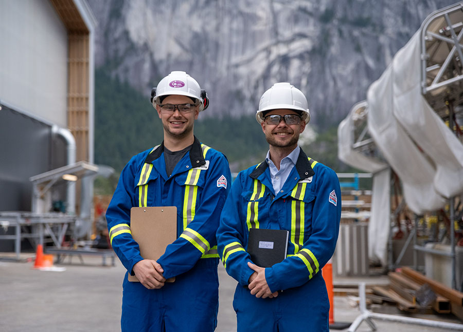 Two Carbon Engineering employees in blue coveralls take measurements outside at a direct air capture facility with mountains in the background and liquid nitrogen in the foreground.